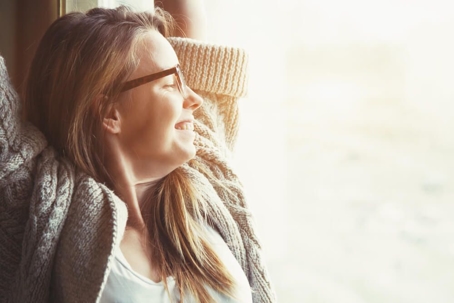A woman wearing glasses and a cardigan is smiling as she looks out the window with her arms raised