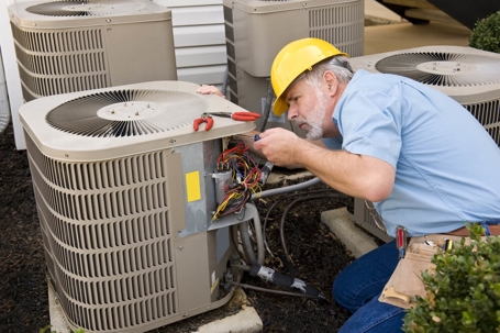 An HVAC technician in a sky blue shirt and denim jeans wearing a khaki tool belt and a yellow hat is working on a central AC unit located outdoors